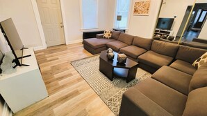 Ein modernes Wohnzimmer mit einem Ecksofa, einem Couchtisch und einem gemusterten Teppich. An der Wand hängt ein großer Spiegel und eine Tür mit einem Glasfenster im Buntglasstil.