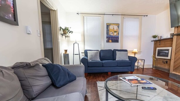 A living room with a grey sofa, a blue sofa, a glass coffee table, and a poster on the wall. - Charming 1-BR Apartment Near Amazing Views in Pittsburgh! (Pittsburgh)