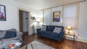 A living room with a grey sofa, a blue sofa, a glass coffee table, and a lamp on a side table. - Charming 1-BR Apartment Near Amazing Views in Pittsburgh! (Pittsburgh)