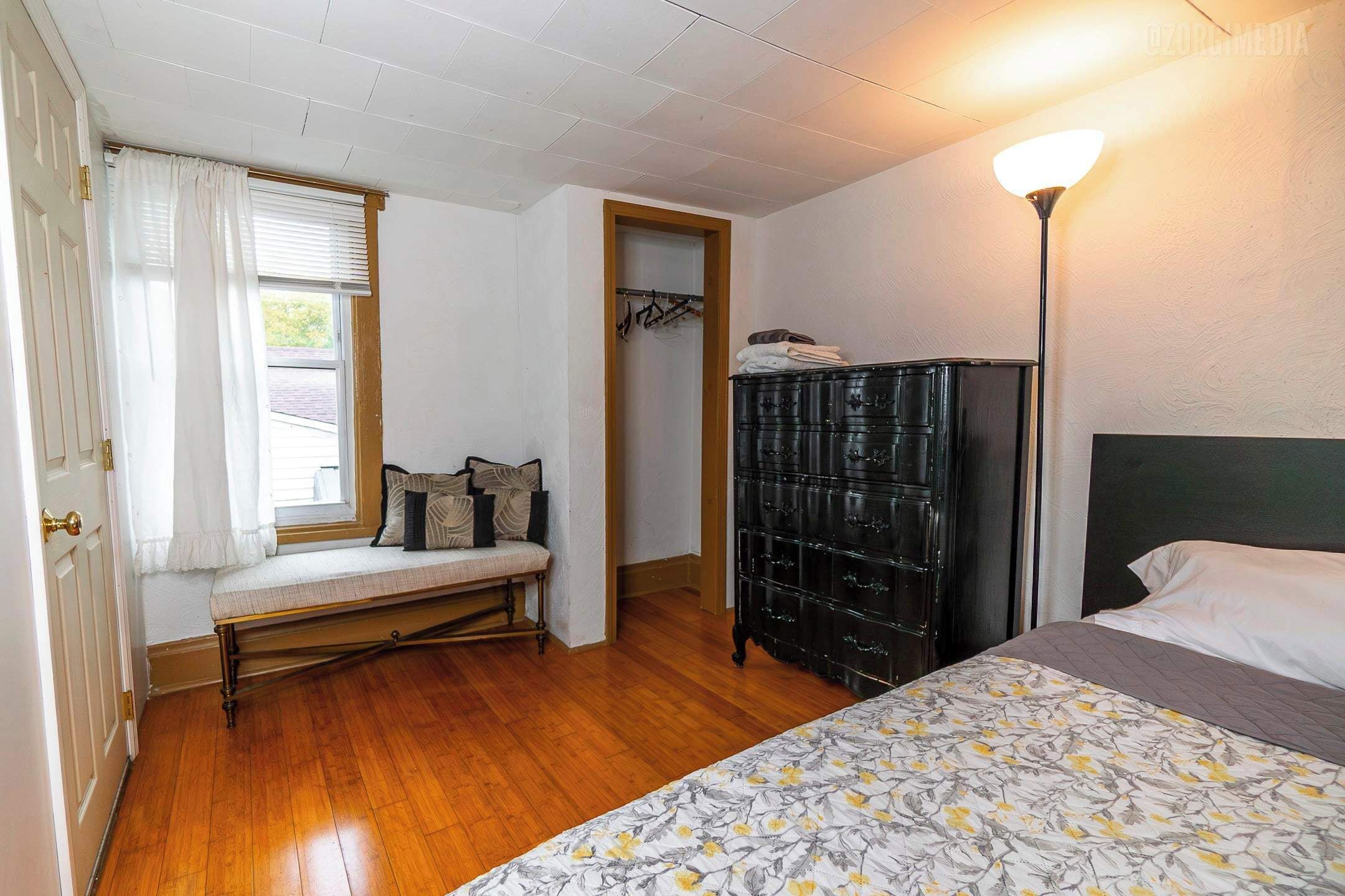 Ein Schlafzimmer mit Holzboden, einer Bank am Fenster, einem Kleiderschrank und einem Bett mit Blumenmusterdecke.