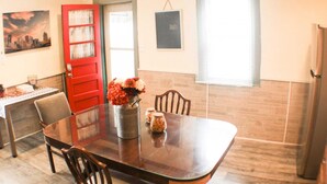 Un comedor con una mesa de madera, sillas, un florero con flores, una puerta roja, una pintura de un paisaje urbano y una pizarra en la pared.