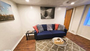 A living room with a dark blue sofa, a small side table, a glass coffee table with decorative items, a patterned rug, and a framed abstract painting on the wall. - Carey Way Quarters - Explorer's Dream Pad (Pittsburgh)