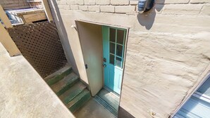 Eine schmale Treppe führt zu einer Tür mit grünem Rahmen, einem Metalllüftungsgitter an der Wand und einer Backsteinmauer.