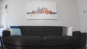 A modern living room with a dark-colored sofa, a cityscape painting on the wall, and a wooden floor. - California Ave Quarters - Modern 1BR - EZ Parking (Pittsburgh)