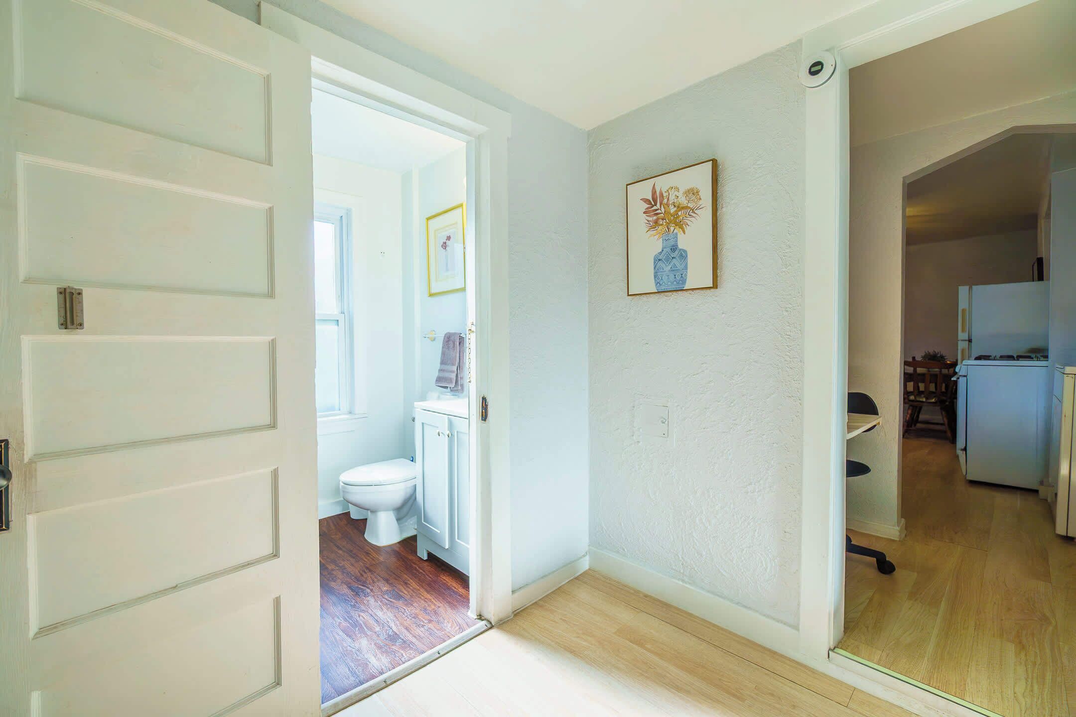 Un pasillo que conduce a un baño con inodoro y armario, un cuadro en la pared, y una cocina visible a través de una puerta abierta.