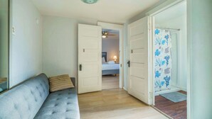 Un couloir avec un canapé gris, un lit à l’arrière-plan et une douche avec un rideau orné de motifs de feuilles.