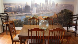 Een eethoek met een tafel en stoelen, een groot raam met uitzicht op de stad, en een decoratieve glazen vaas op de tafel.