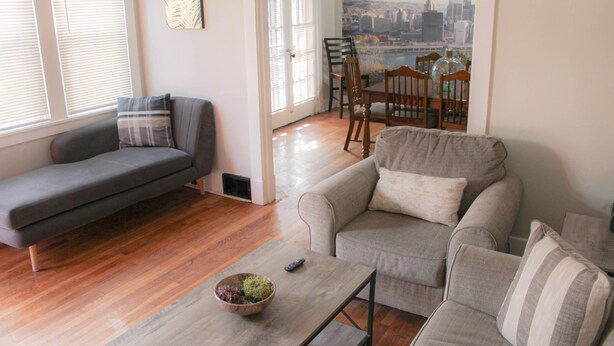 Een woonkamer met een grijze bank, een grijze fauteuil, een houten eettafel en een stadsgezicht op de muur.
