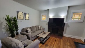 Een woonkamer met een grijze bank, een salontafel en een open haard. Er staan twee potplanten en een flatscreen televisie is aan de muur bevestigd.