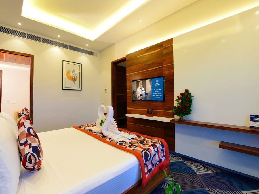 Habitación de hotel con una cama bien hecha, un televisor montado en la pared, un mostrador de recepción y un arreglo floral.