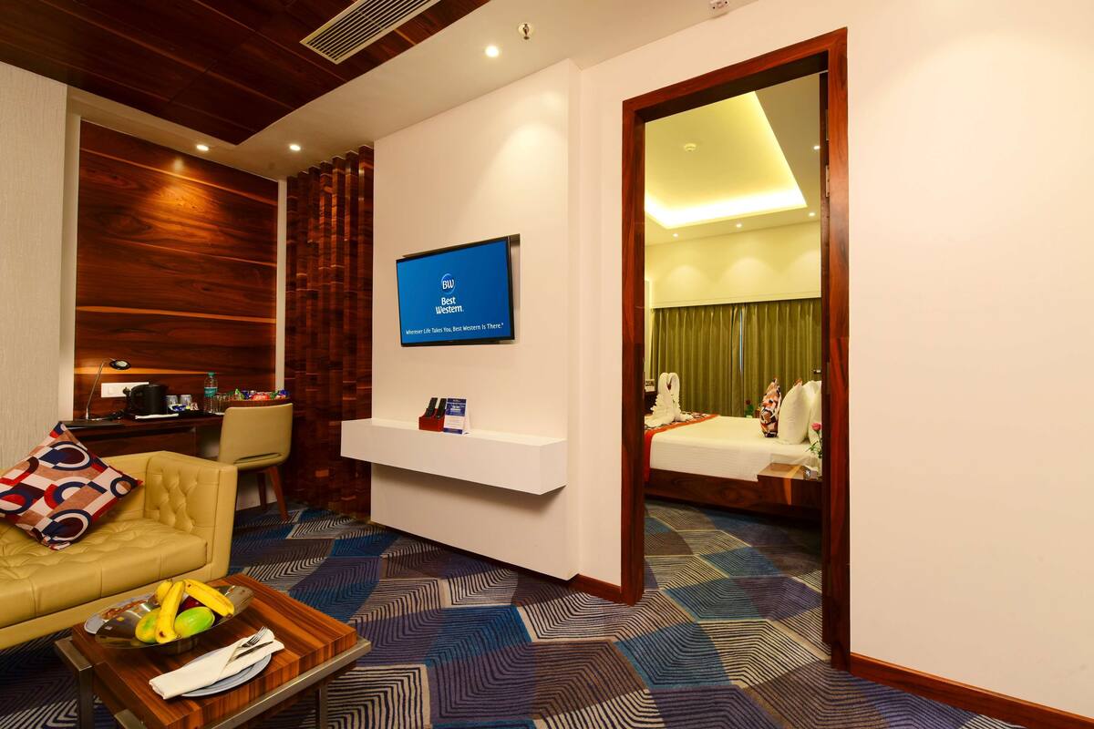 Een hotelkamer met een houten accentmuur, een aan de muur bevestigde televisie, een bureau met stoel, een bank met een gekleurd kussen, een dienblad met fruit en een servet, en een bed met een groen gordijn.