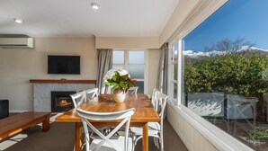 A dining area with a wooden table and white chairs, a fireplace, and a large window with a view of a snowy mountain. - Townside Treat - Wanaka Holiday Home (Wanaka)