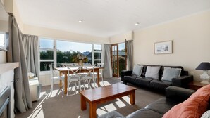 A spacious living room with a wooden coffee table, a black leather sofa, and a dining area with white chairs and a table. - Townside Treat - Wanaka Holiday Home (Wanaka)