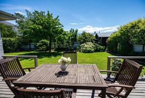 A wooden deck with a table and chairs, a vase with flowers, and a view of a well-maintained garden and house in the background.