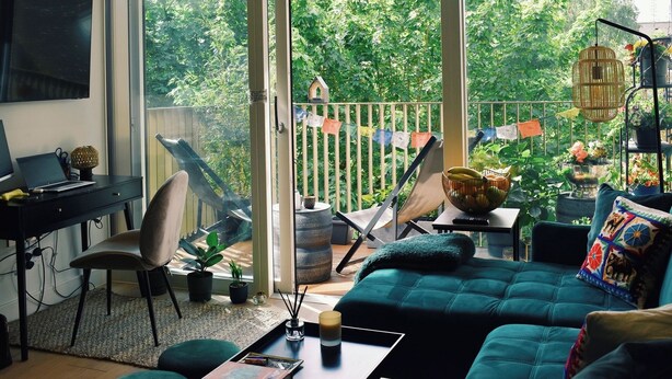 A modern living room with a large window, a desk with a laptop, a green sofa, and a balcony with plants and a hanging chair. - Urban Oasis | 1Bed | Pvt Balcony | Westbourne Park (London)