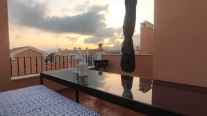 A balcony with a glass table, a patterned cushion, and a view of buildings and a sunset. - Villa Terreros 365 days of sunshine (Pulpí)