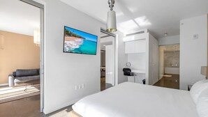 Ein modernes Hotelzimmer mit einem großen Bett, einem flachbildfernseher an der Wand, einem Schreibtisch mit Stuhl und einem durchgehenden Türspalt einsehbaren Badezimmer.