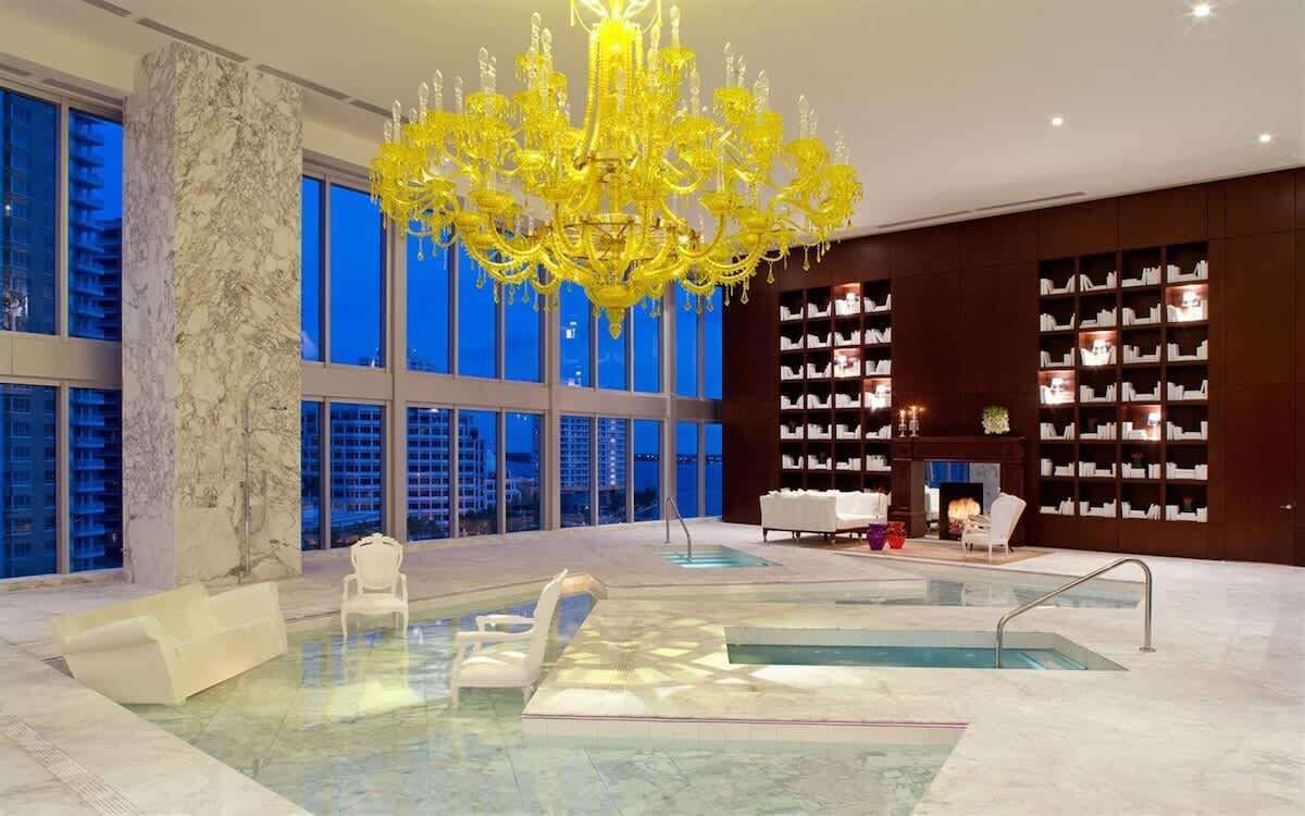 Un área de piscina interior lujosa con una gran lámpara de araña, columnas de mármol y vistas a la ciudad.
