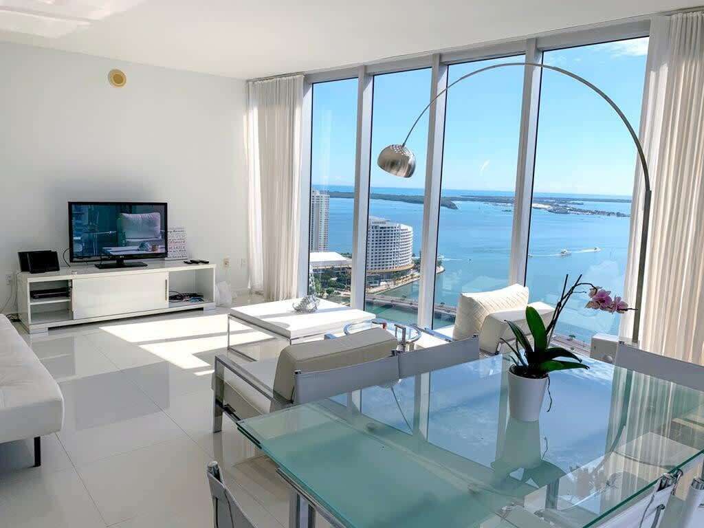 Una sala de estar moderna con un ventanal grande, un televisor de pantalla plana, muebles blancos y una mesa de comedor de vidrio.