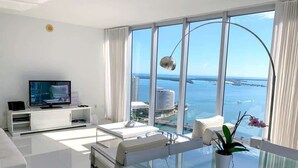 A modern living room with a large window, a flat-screen TV, white furniture, and a glass dining table. - Icon Brickell Condo with Bay Views (Miami)