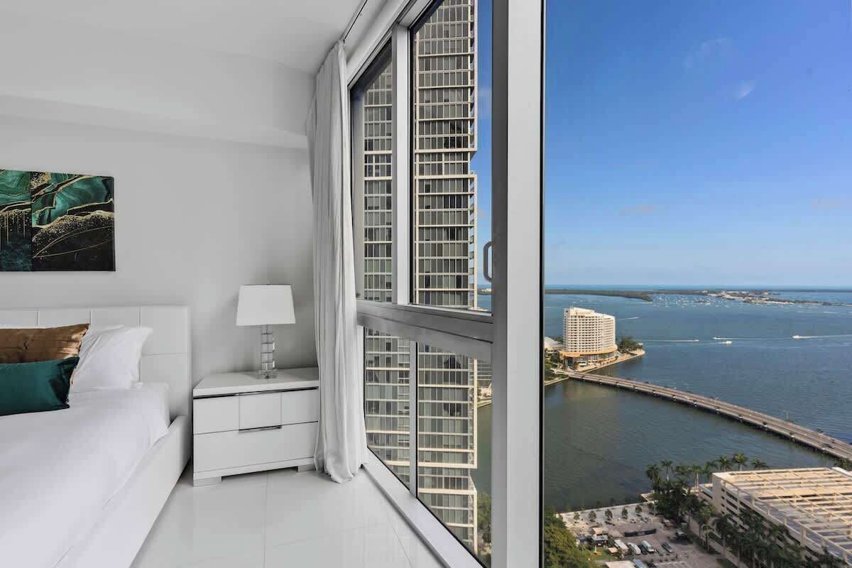 A hotel room with a large window overlooking a coastal cityscape, a bed, a nightstand, and a painting on the wall.