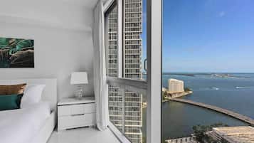 A hotel room with a large window overlooking a coastal cityscape, a bed, a nightstand, and a painting on the wall.