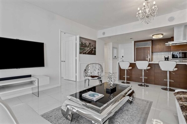 A modern living room with a flat-screen TV, a glass coffee table, a chandelier, and a kitchen area with bar stools.