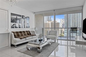 Un salón moderno con un sofá blanco, una mesa de centro de vidrio y vistas a la ciudad a través de amplias ventanas.