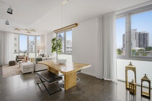A modern living room with a wooden dining table, a sofa set, and a large window with curtains.