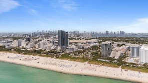 Un paisaje urbano costero con edificios altos, una playa con tumbonas y un cielo despejado.