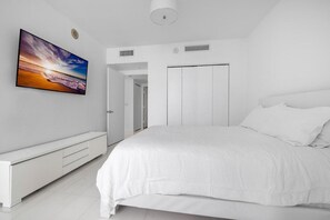 A bedroom with a large bed, a white wardrobe, a flat-screen TV mounted on the wall, and a white bench.