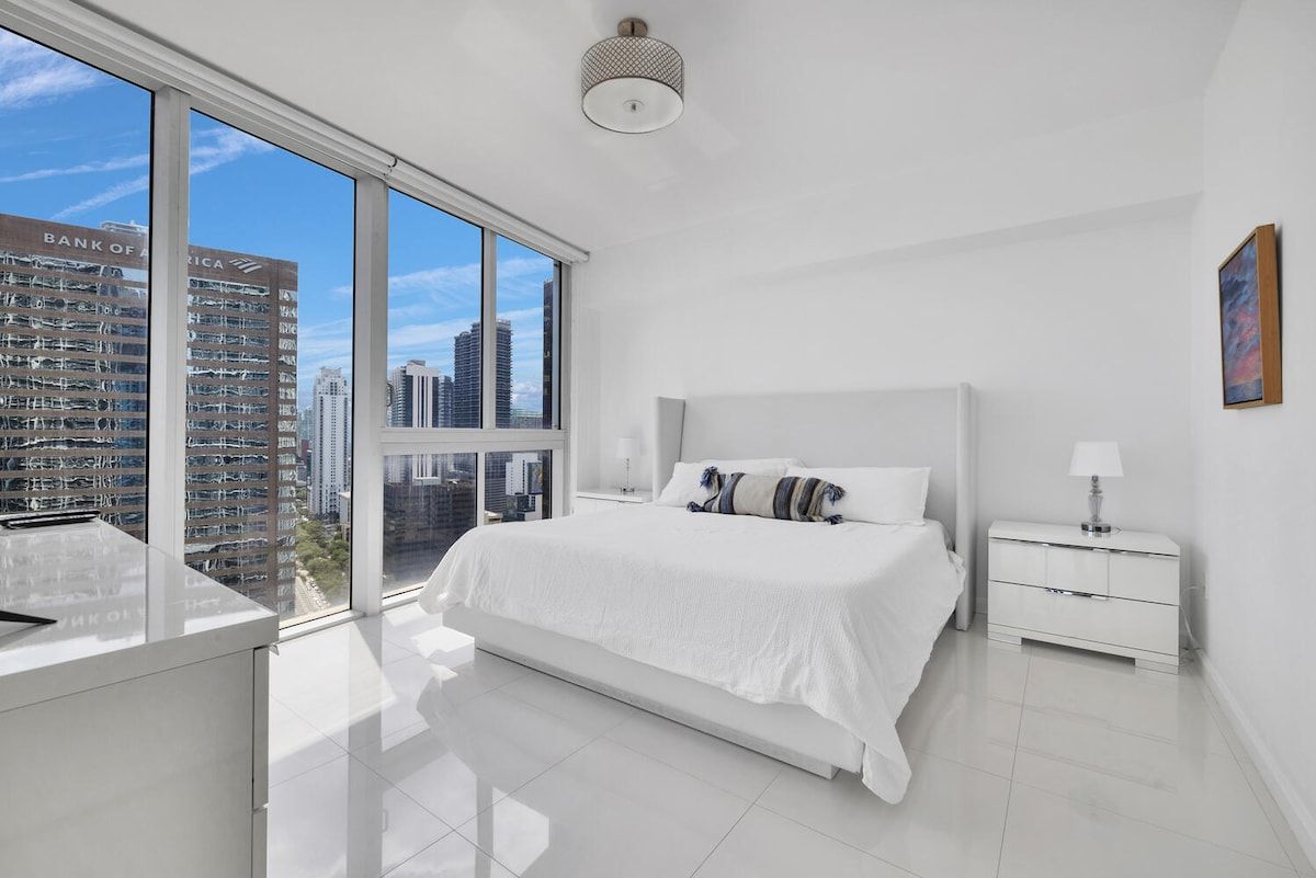 A modern bedroom with a large bed, a bedside table, a nightstand, and a view of a cityscape through floor-to-ceiling windows.
