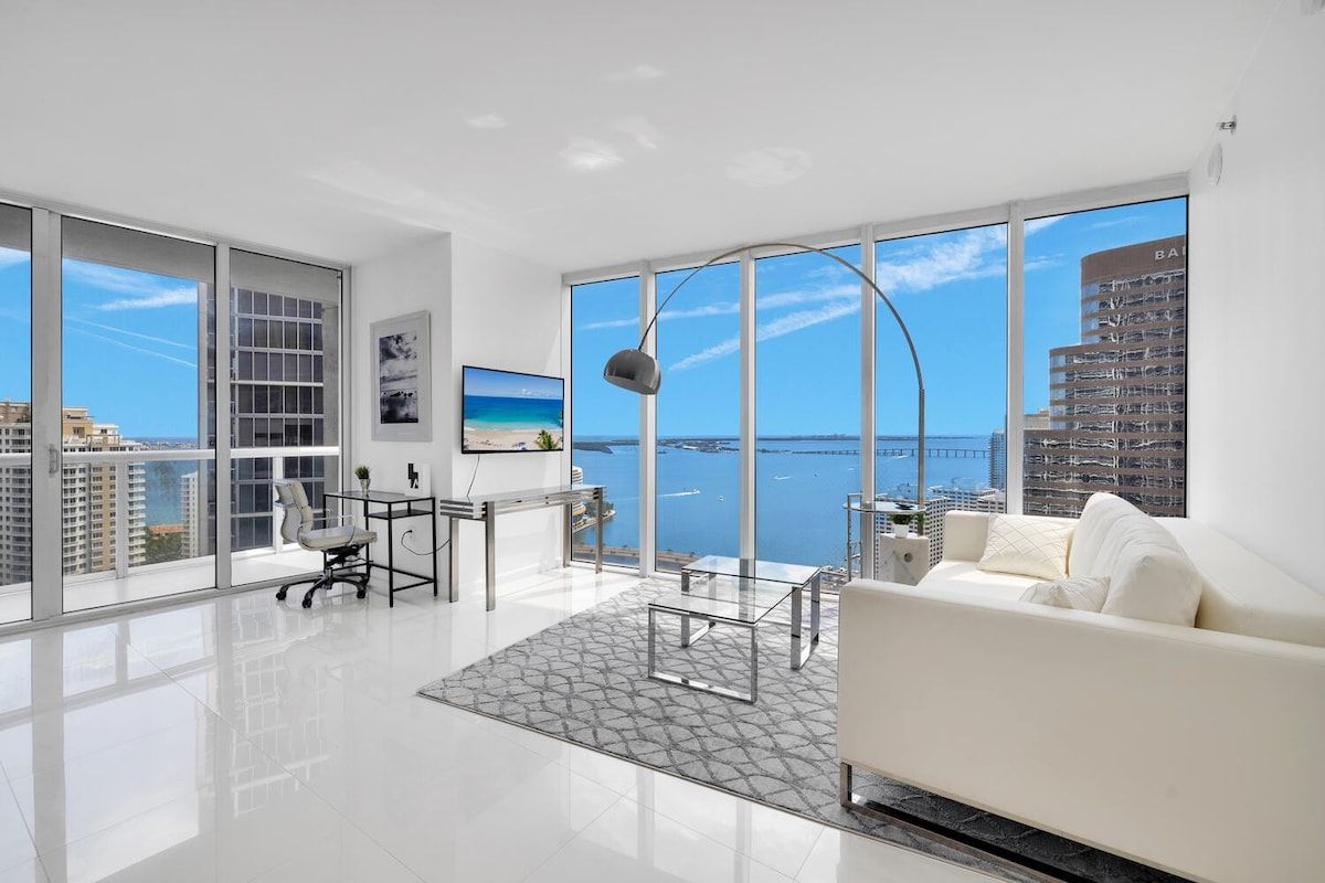 A modern living room with a white sofa, a glass coffee table, and a desk with a chair. Large windows offer a view of the sea and buildings.