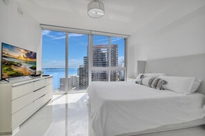 A modern bedroom with a large bed, a TV on a dresser, and a view of a cityscape through floor-to-ceiling windows.