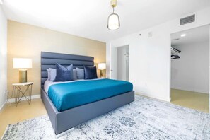 Una habitación de hotel moderna con una cama grande, lámparas de noche, una alfombra con diseño de mármol y una puerta que da a un pasillo.