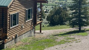 A log cabin with a metal roof, large windows, and a satellite dish. It is situated on a grassy hill with a view of distant mountains and a clear sky. - Moonlight Ridge Cabin (Ennis)