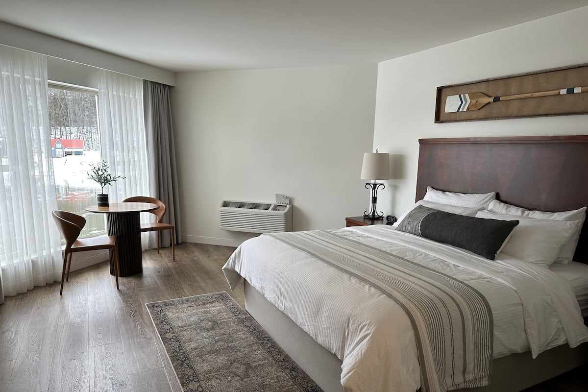 A hotel room with a bed, a bedside table with a lamp, a chair, a small table with a plant, and a view of the outside through large windows.