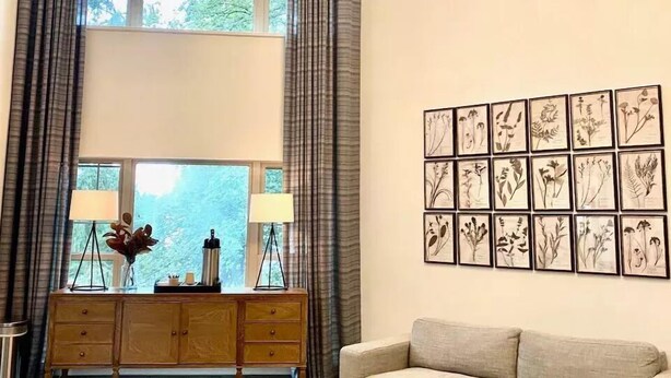 A modern living room with a large window, a chandelier, a sofa, a coffee table, and a wall of framed botanical prints. - Foxburg Inn (Foxburg)