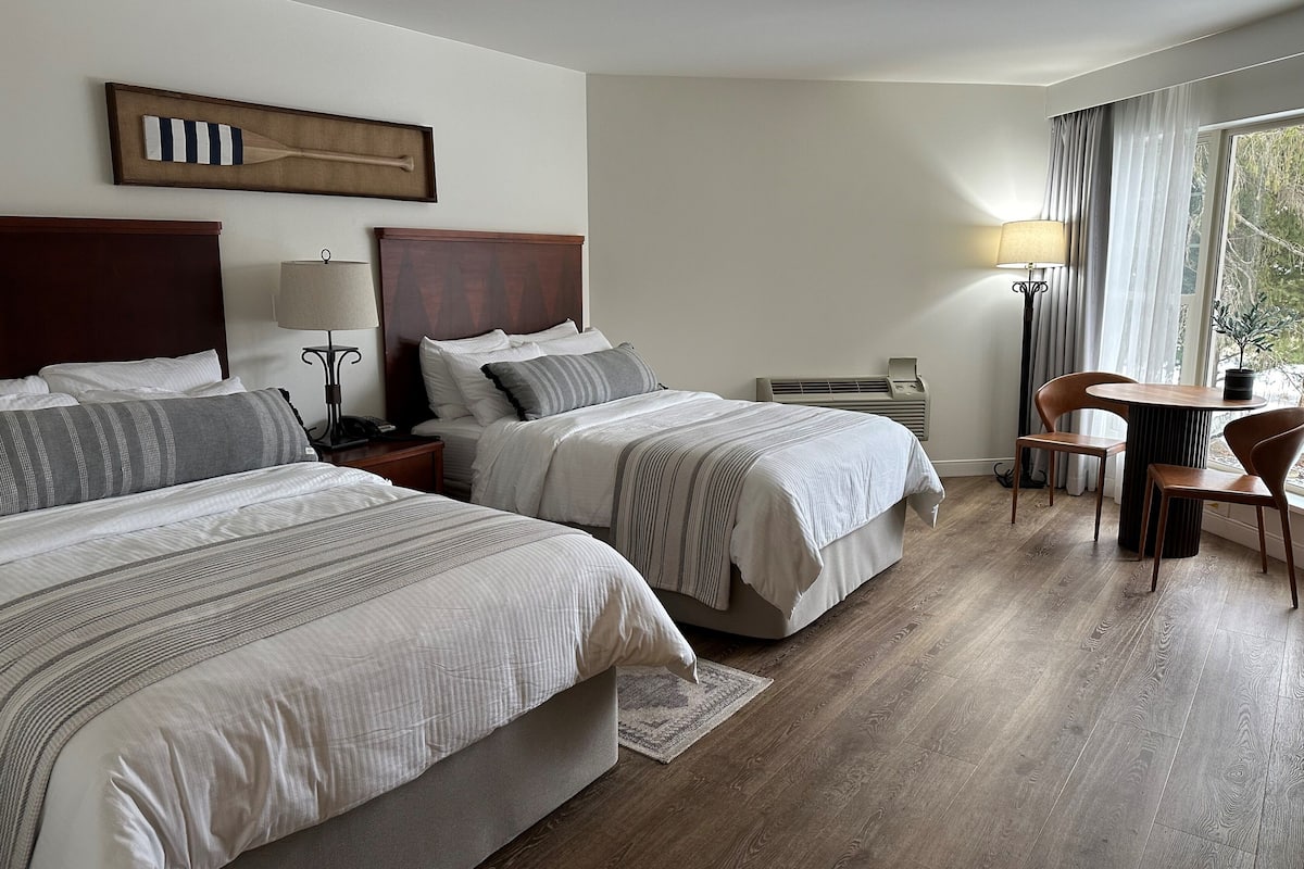 A hotel room with two beds, a bedside table with a lamp, a small table with chairs, and a window with curtains.