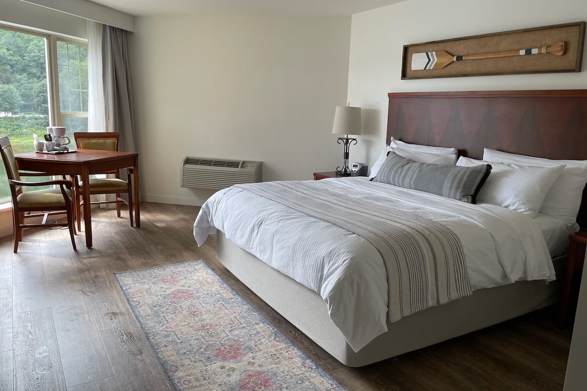 A hotel room with a large bed, a bedside table with a lamp, a chair, a table with cups and a coffee pot, and a view of greenery through the window.