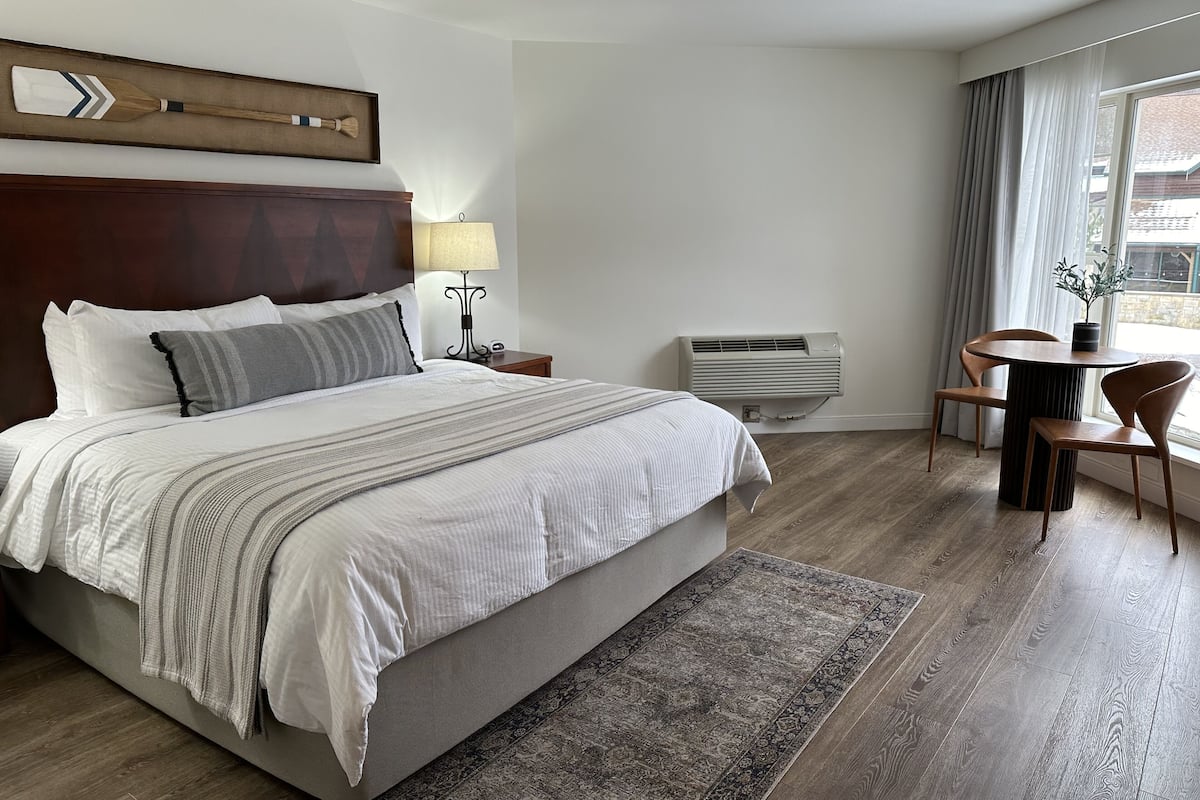 A hotel room with a large bed, a bedside table with a lamp, a small table with chairs, and an air conditioning unit.