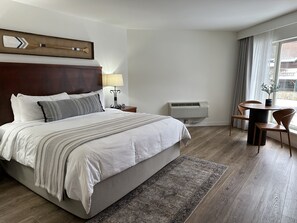 A hotel room with a large bed, a bedside table with a lamp, a small table with chairs, and an air conditioning unit.