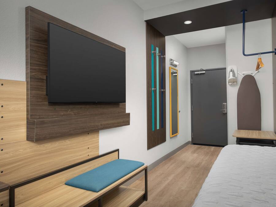 Una camera d'albergo moderna con un letto grande, una panca con un cuscino blu, una televisione a schermo piatto montata a parete, un armadio con specchio e una porta con maniglia.