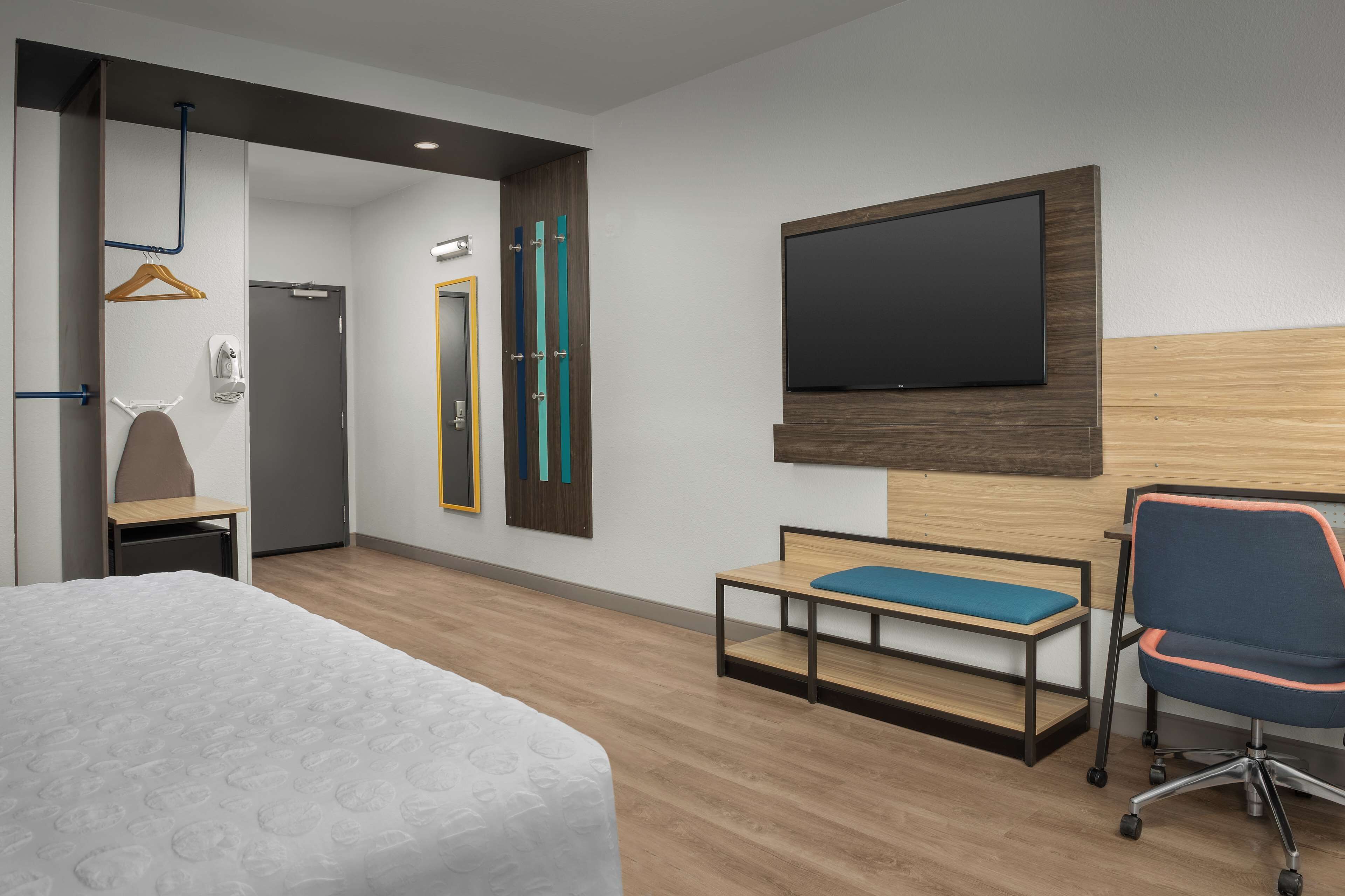 A hotel room with a bed, a desk, a chair, a TV, and a bench.
