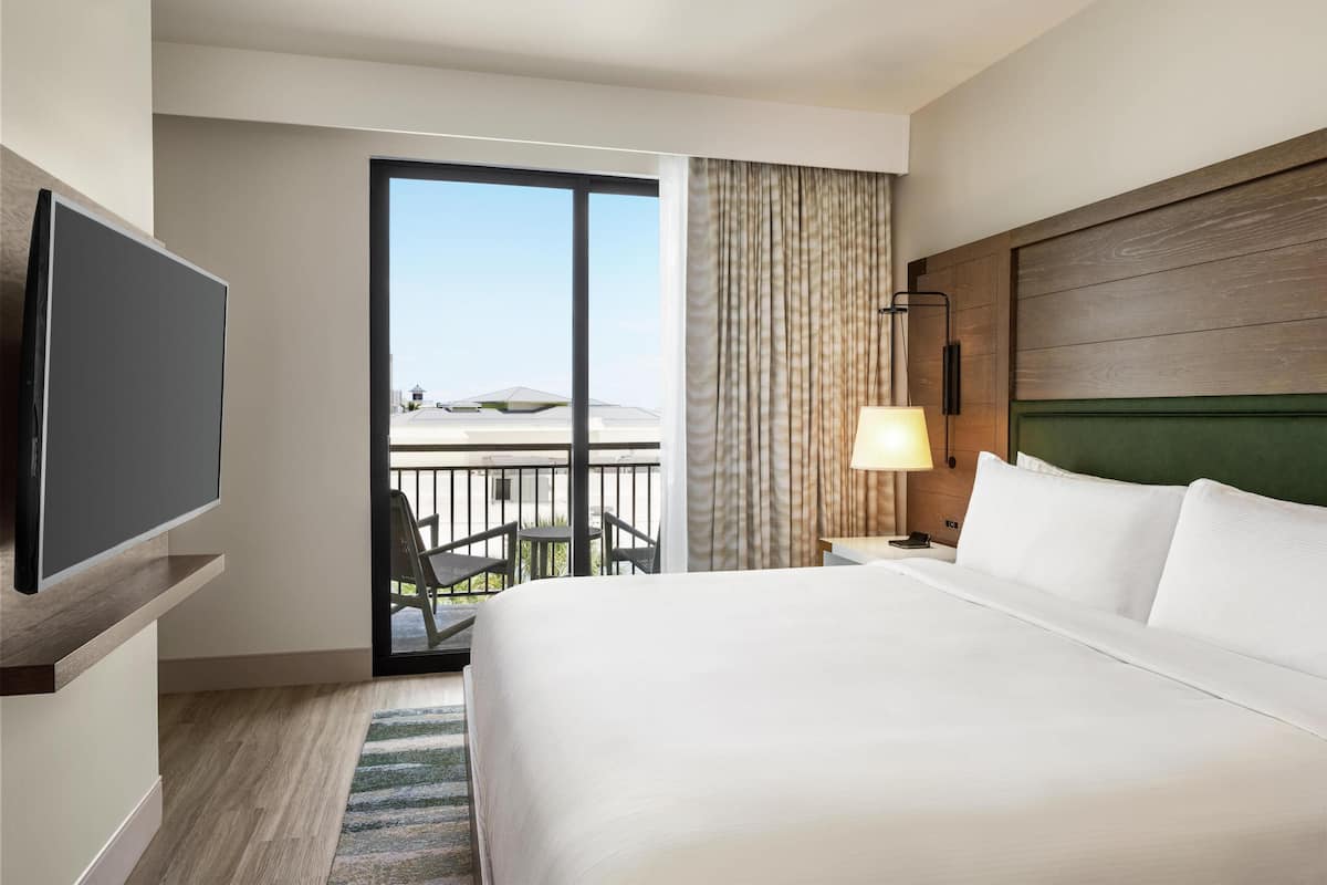 A hotel room with a large bed, a flat-screen TV, a balcony with a view, and a modern headboard.