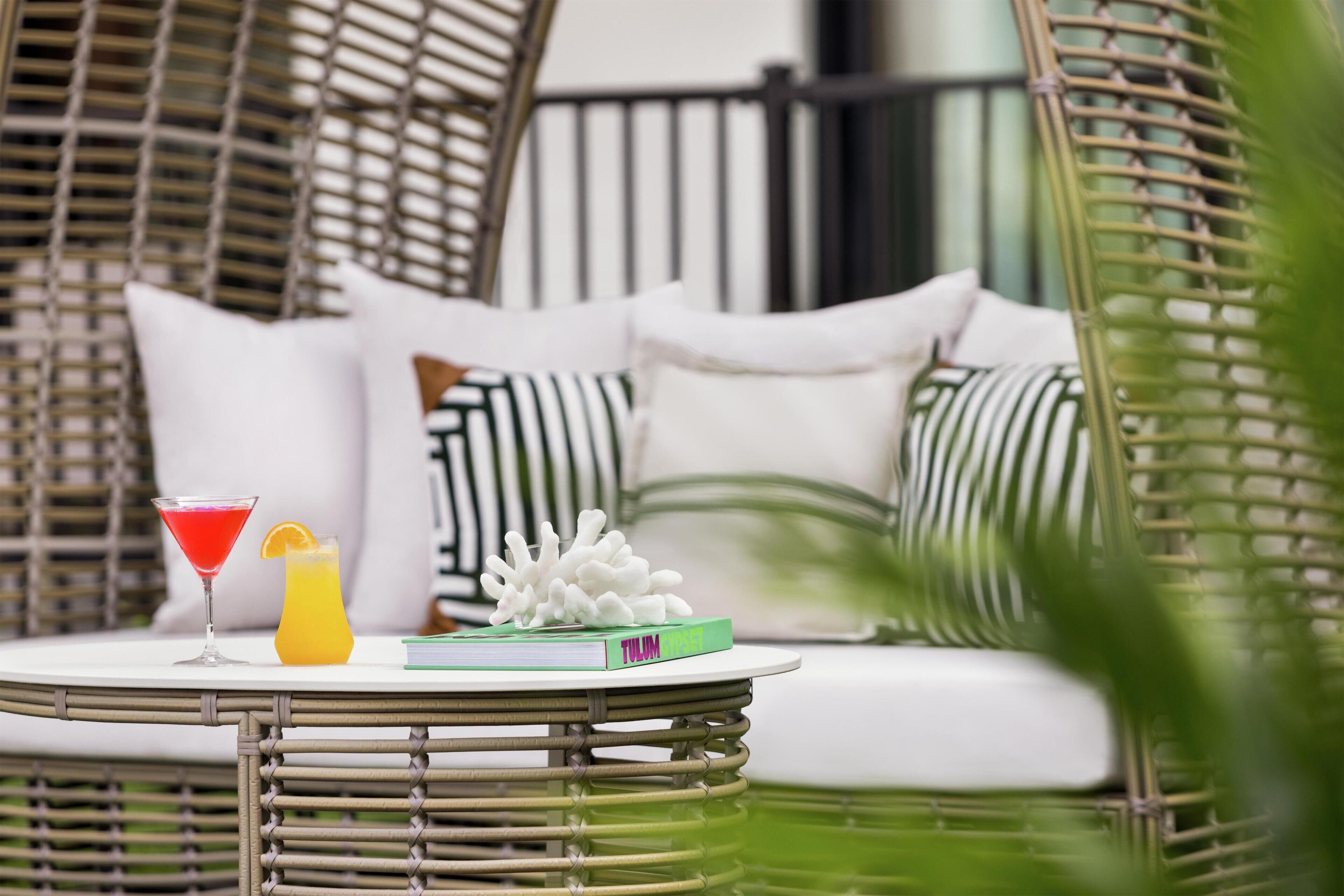 Una silla de mimbre con cojines blancos, una mesita redonda con un libro y bebidas, y una planta verde al frente.