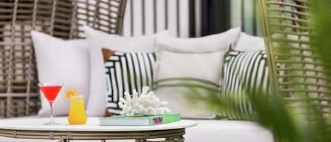 Una silla de mimbre con cojines blancos, una mesita redonda con un libro y bebidas, y una planta verde al frente.