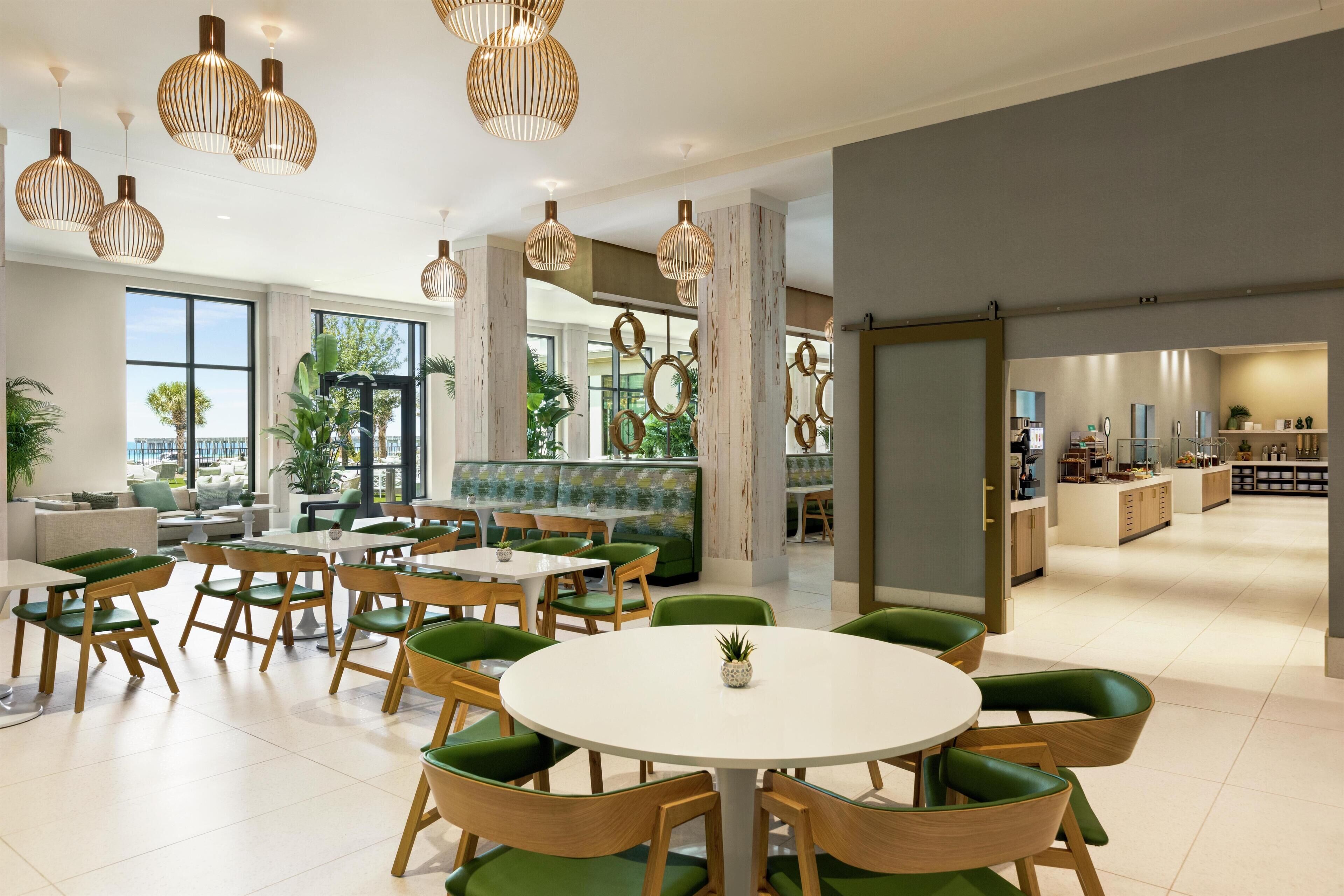 A modern dining area with round tables and green chairs, large windows, and a view of the outdoors.