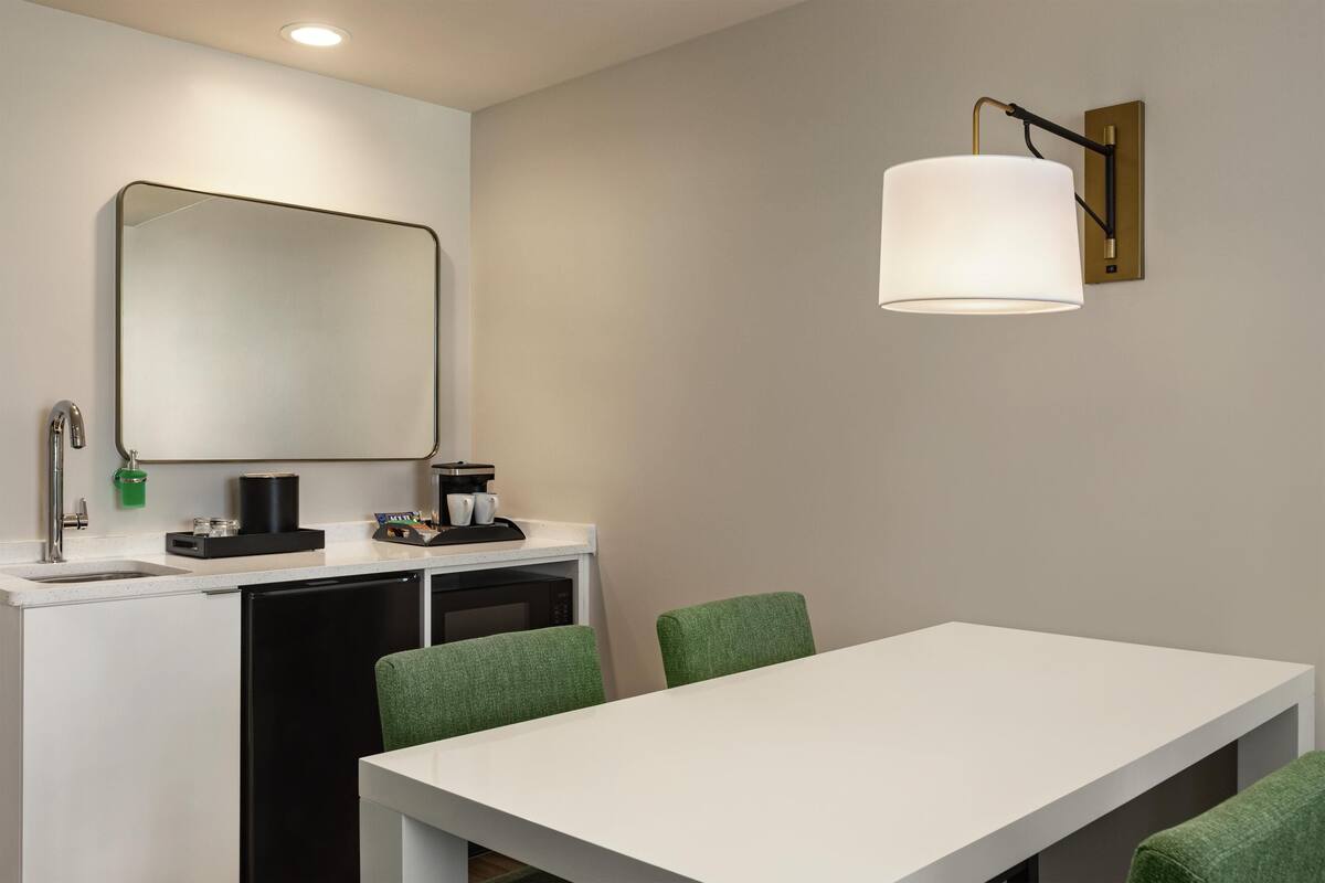 A modern hotel room with a white dining table, green upholstered chairs, a built-in microwave, and a large mirror above the sink.