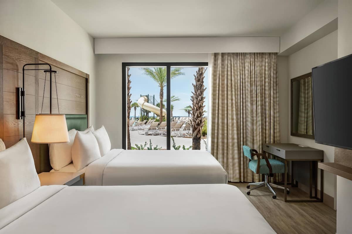 A hotel room with a bed, a desk, a chair, a TV, and a view of a pool and palm trees.